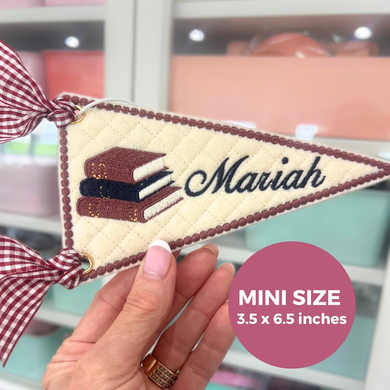 May include: A cream-colored quilted triangular pennant featuring an embroidered stack of three books and the name Mariah in dark blue script. The pennant has a dark red and white gingham ribbon attached and measures 3.5 x 6.5 inches.