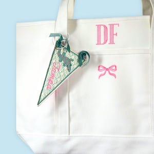 May include: A white canvas tote bag featuring the embroidered pink letters DF and a pink bow. A triangular pennant tag hangs from the handle, displaying a green lizard illustration, a floral pattern, and the pink embroidered text Tampa.