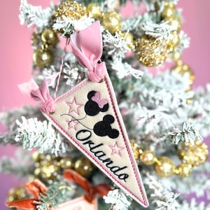 May include: A cream-colored pennant-shaped Christmas ornament with the word "Orlando" in black script. The ornament features black Mickey Mouse silhouettes, pink stars, and a pink ribbon for hanging. The ornament is displayed on a flocked Christmas tree.
