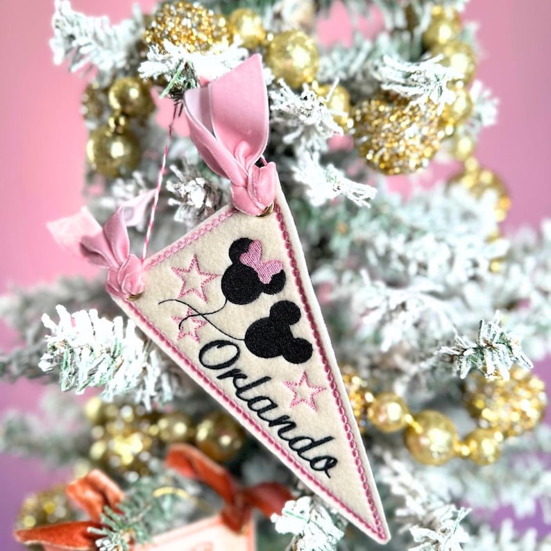 May include: A cream-colored pennant-shaped Christmas ornament with the word "Orlando" in black script. The ornament features black Mickey Mouse silhouettes, pink stars, and a pink ribbon for hanging. The ornament is displayed on a flocked Christmas tree.