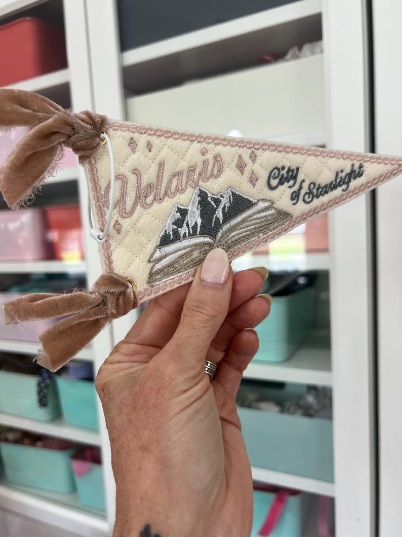 May include: A triangular cream-colored quilted pennant featuring embroidered text that reads Velaris City of Starlight. The design includes a central open book with dark gray mountain peaks rising from the pages and two brown velvet ribbons.