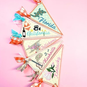 May include: Five triangular fabric pennants featuring embroidered designs and text. The pennants display the words Florida, California, Tullahoma, Nashville, and Kentucky, accompanied by icons like a lizard, bikini, airplane, guitar, and flower.