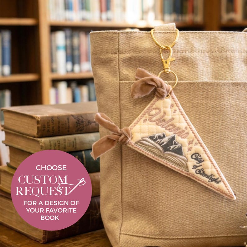 May include: A beige canvas tote bag featuring a triangular quilted fabric keychain. The keychain displays embroidered text reading Velaris City of Starlight above an open book illustration with mountains, accented by two light brown velvet ribbons.