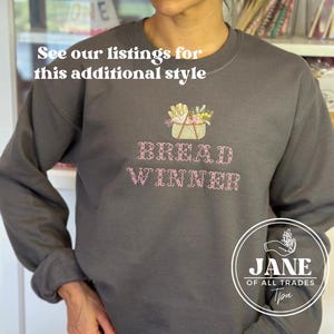 May include: A gray sweatshirt with a pink embroidered design of a basket of bread and the text "BREAD WINNER". The sweatshirt has a logo on the bottom right corner that says "JANE OF ALL TRADES".