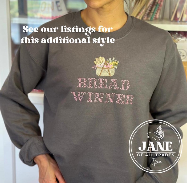 May include: A gray sweatshirt with a pink embroidered design of a basket of bread and the text "BREAD WINNER". The sweatshirt has a logo on the bottom right corner that says "JANE OF ALL TRADES".