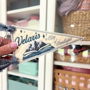 May include: A triangular cream-colored felt pennant featuring quilted stitching and dark blue embroidery. The text reads Velaris City of Starlight above an open book with mountain peaks. The pennant has frayed blue fabric ties at the corners.