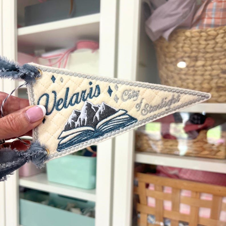 May include: A triangular cream-colored felt pennant featuring quilted stitching and dark blue embroidery. The text reads Velaris City of Starlight above an open book with mountain peaks. The pennant has frayed blue fabric ties at the corners.