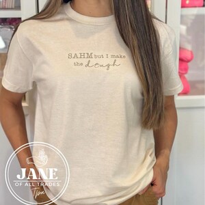 May include: A white t-shirt with the text "SAHM but I make the dough" printed on the front. The shirt is being worn by a person.