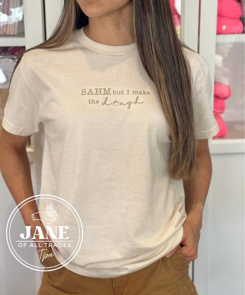 May include: A white t-shirt with the text "SAHM but I make the dough" printed on the front. The shirt is being worn by a person.