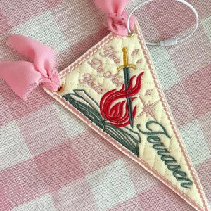 May include: A triangular cream-colored quilted pennant featuring embroidered designs of an open book, a sword, and flames. The text reads "You Do Not Yield" and "Terrasen" in dark green script. It is accented with pink chiffon ribbons.