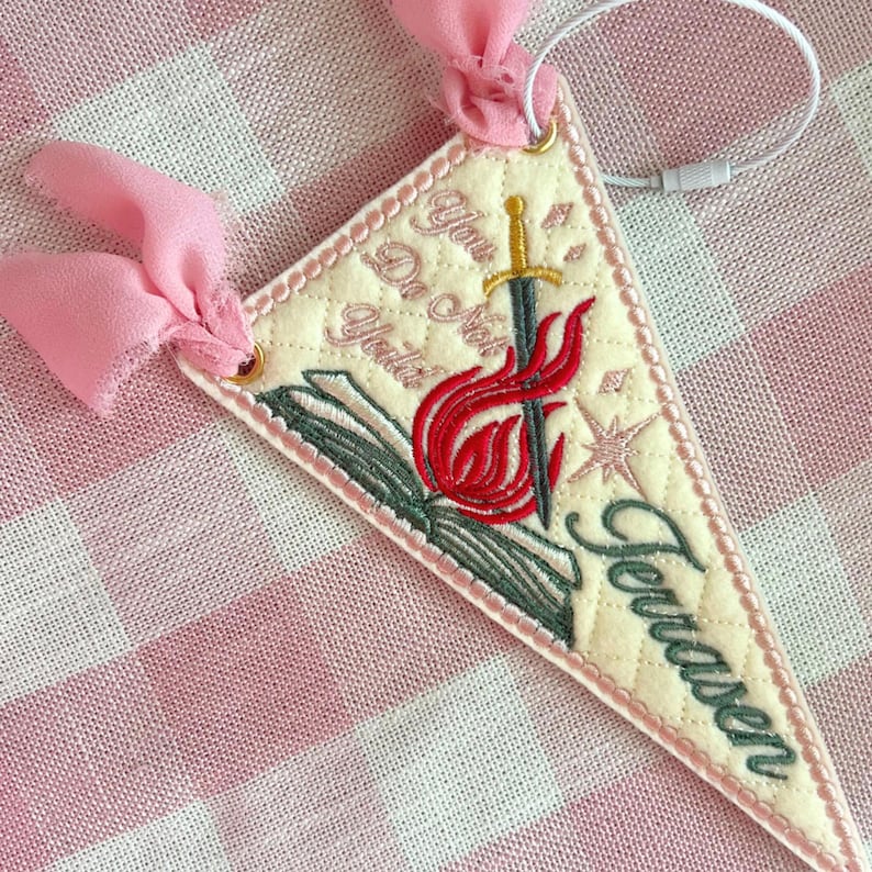 May include: A triangular cream-colored quilted pennant featuring embroidered designs of an open book, a sword, and flames. The text reads "You Do Not Yield" and "Terrasen" in dark green script. It is accented with pink chiffon ribbons.