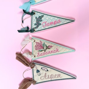 May include: Mini felt pennant city ornaments. Each triangular pennant features embroidered designs and city names like Miami, Tampa, Savannah, and Aspen. They have decorative trim and are adorned with ribbons.