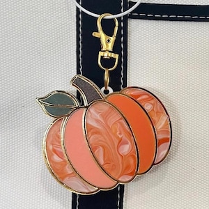 Könnte beinhalten: Ein kürbisförmiger Schlüsselanhänger mit goldfarbenem Metallrahmen und Karabinerverschluss. Der Kürbis hat eine marmorierte orange-weiße Acrylfüllung mit einem dunkelgrünen Blattakzent oben, befestigt an einem schwarzen Stoffband.