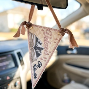 May include: A triangular quilted fabric car ornament in cream with a brown velvet ribbon hanger. It features an embroidered open book with mountain peaks, small star accents, and the text "Velaris City of Starlight" in dark brown thread.
