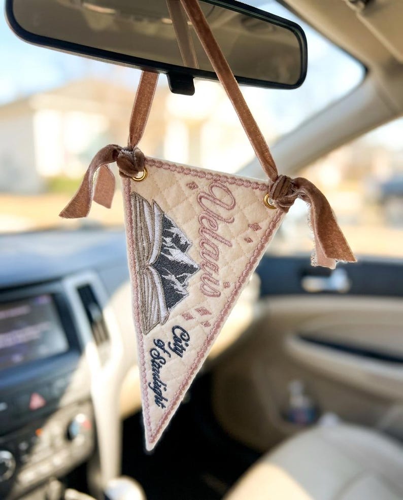 May include: A triangular quilted fabric car ornament in cream with a brown velvet ribbon hanger. It features an embroidered open book with mountain peaks, small star accents, and the text "Velaris City of Starlight" in dark brown thread.