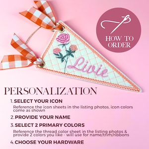 May include: A triangular fabric pennant featuring an embroidered design of three pink roses and the name Livie in pink script. The pennant has a quilted diamond pattern, orange gingham ribbons, and metal grommets. Text includes HOW TO ORDER and PERSONALIZATION.