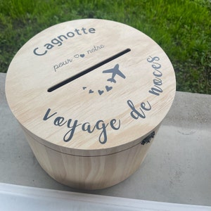 Peut inclure: Une boîte en bois avec une fente sur le dessus pour l'argent. La boîte est décorée du texte "Cagnotte pour notre voyage de noces" et d'un petit avion avec des cœurs.