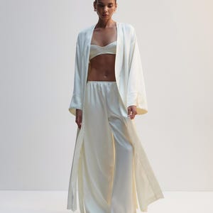 Puede incluir: Un conjunto de tres piezas en satén blanco roto que consta de una bata abierta de manga larga con puños anchos, un top bralette corto con escote corazón y pantalones de talle alto y pernera ancha con un acabado fluido y suave.