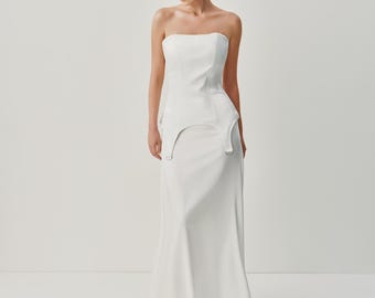 May include: A floor-length white strapless gown featuring a structured corset-style bodice with a peplum waist detail. The garment includes two silver-toned D-ring hardware accents at the front hips and a smooth, flowing skirt that flares slightly.