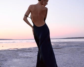 May include: A floor-length black slip dress featuring a deep scoop back, thin spaghetti straps, and a fluid, bias-cut silhouette that drapes down to the ground. The garment is made of a smooth, dark fabric with a subtle sheen.