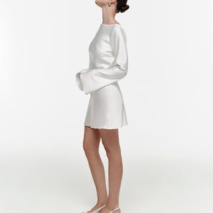 Puede incluir: Un vestido blanco de manga larga y acampanada, con cuello barco. El vestido se combina con zapatos de tacón blancos con correa trasera. La modelo mira hacia arriba. El fondo es blanco.