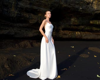 May include: A white, floor-length wedding dress with a simple, elegant design. The dress features a fitted bodice, spaghetti straps, and a flowing skirt with a slight train. The model is standing on a black sandy beach with a dark rock formation in the background.
