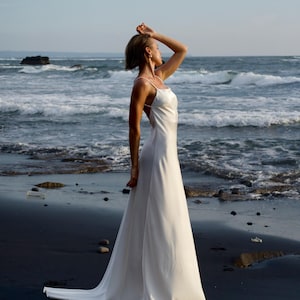 May include: A white satin wedding dress with spaghetti straps and a flowing A-line skirt. The dress is worn on a beach with dark sand and the ocean in the background. The dress has a low back and a slight train.