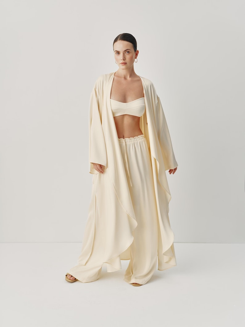 May include: A white long-sleeved robe with a draped design and wide-leg pants. The robe has a relaxed fit and a shawl collar. The pants have a drawstring waist and a wide leg.