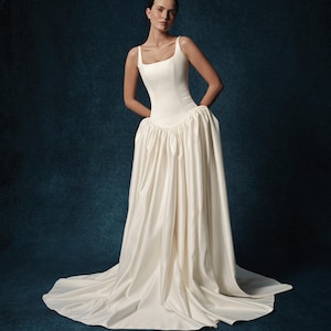 May include: A floor-length ivory gown featuring a structured, fitted bodice with a square neckline and thick straps. The waistline drops into a voluminous, pleated skirt that flows to the floor, creating a full silhouette with a train.