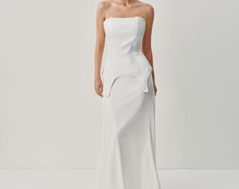 May include: A floor-length white strapless gown featuring a structured corset-style bodice with a peplum waistline. The bodice includes two silver-toned metal D-rings attached to the front hem, transitioning into a smooth, flowing skirt.
