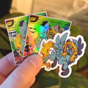 Könnte beinhalten: Eine Hand hält mehrere Aufkleber mit Cartoon-Designs. Die Aufkleber zeigen ein blaues und gelbes geflügeltes Pony, ein Trollgesicht und andere Grafiken auf einem grünen und holografischen Hintergrund. Die Aufkleber haben ein glänzendes Finish.