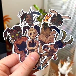 May include: A selection of stickers showcasing figures with bunny ears and diverse attire. The stickers have a glittery finish and a dark, slightly eerie aesthetic. The figures are shown in various poses and expressions, creating a unique collection.