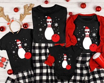 May include: Black Christmas-themed shirts and a onesie featuring a bowling pin wearing sunglasses and a Santa hat, with a red bowling ball. The shirts are paired with black and white plaid pajama pants. Festive holiday apparel.