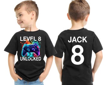 Könnte beinhalten: Zwei schwarze T-Shirts für Kinder. Ein Shirt hat ein buntes Videospiel-Controller-Motiv mit dem Text "Level 8 Unlocked". Das andere Shirt hat den Text "Jack 8" in Weiß.