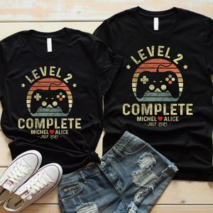 May include: Two black t-shirts with a retro video game controller graphic and the text "Level 2 Complete" with a heart and names. The text below the graphic says "July 2021".