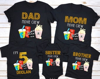 May include: Black t-shirts with movie-themed graphics. Designs include "DAD MOVIE CREW," "MOM MOVIE CREW," "I'M 5 DECLAN," "SISTER MOVIE CREW," and "BROTHER MOVIE CREW." Each design features popcorn, a drink, film clapper, and movie tickets.
