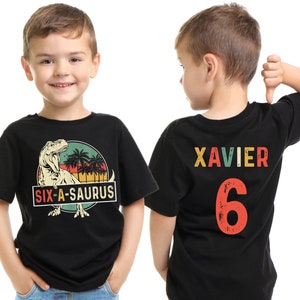May include: Black t-shirt with a dinosaur graphic and the text "SIX-A-SAURUS" on the front. The back of the shirt displays the name "XAVIER" and the number "6" in colorful letters. The shirt is designed for a child.