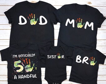 May include: A set of five black t-shirts with colorful handprint designs and text. The shirts say "DAD", "MOM", "SISTR", "BR", and "I'm officially 5 Olivia a handful".