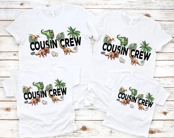May include: Four white t-shirts with a dinosaur and palm tree design. The shirts have the text "Cousin Crew" printed on them.