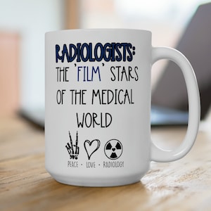 Pode incluir: Caneca de café de cerâmica branca com o texto "Radiologists: The 'Film' Stars Of The Medical World" em letras azuis. Abaixo do texto, há três ícones: um sinal de paz, um coração e um símbolo de radiação. O texto "Peace - Love - Radiology" está abaixo dos ícones.