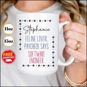 Pode incluir: Caneca de cerâmica branca com uma borda de pegadas de pata pretas. O texto "Stephanie Feline Lover. Paycheck Says Software Engineer" está impresso na caneca.
