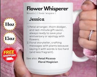 May include: White ceramic mug with a black and white design that reads "Flower Whisperer Noun(n.) / flavə 'wispərə / Jessica 1. Petal arranger, thorn dodger, and last-minute gift savior, always ready to save your anniversary or apology with flowers. 2. Floral storyteller, crafting messages with plants because saying it with words is too hard (and less fragrant). See also: Petal Picasso Floral Magician" The mug is being held by a hand wearing a pink sweater.