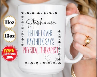 Pode incluir: Caneca de cerâmica branca com pegadas pretas ao redor da borda. A caneca tem texto preto que diz "Stephanie Feline Lover. Paycheck Says Physical Therapist" com um coração vermelho e uma pegada na parte inferior.