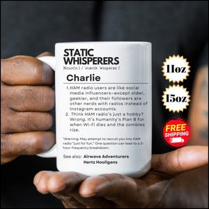 Puede incluir: Taza de cerámica blanca con el texto "STATIC WHISPERERS" y una definición. La taza también presenta el nombre "Charlie" y los tamaños 11oz y 15oz. La taza es sostenida por una persona.