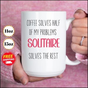 May include: White ceramic mug with the text "COFFEE SOLVES HALF OF MY PROBLEMS SOLITAIRE SOLVES THE REST" in black and pink. The mug is held by a person. The mug is available in 11oz and 15oz sizes.