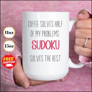 Può includere: Tazza in ceramica bianca con la scritta "COFFEE SOLVES HALF OF MY PROBLEMS SUDOKU SOLVES THE REST" in nero e rosa. La tazza è tenuta da una persona. Disponibile in 312 ml e 425 ml.