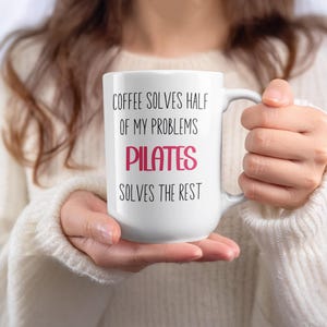 Może przedstawiać: Biały ceramiczny kubek do kawy z napisem "COFFEE SOLVES HALF OF MY PROBLEMS PILATES SOLVES THE REST" w czarnych i różowych literach. Kubek ma duży uchwyt i jest trzymany przez osobę w białym swetrze.