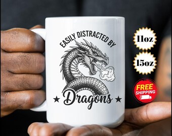 Puede incluir: Taza de cerámica blanca con un diseño de dragón en blanco y negro y el texto "EASILY DISTRACTED BY Dragons". Disponible en tamaños de 325 ml y 444 ml.