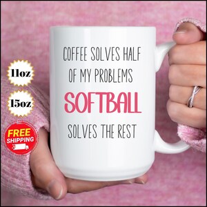 May include: White ceramic mug with the text "COFFEE SOLVES HALF OF MY PROBLEMS SOFTBALL SOLVES THE REST" in black and pink. The mug is held by a person. The mug is available in 11oz and 15oz sizes.