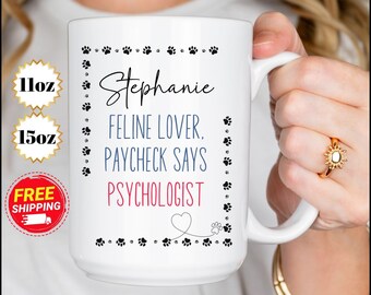 May include: White ceramic mug with black paw prints around the edge. The mug says "Stephanie Feline Lover. Paycheck Says Psychologist" in black and blue text with a pink heart and paw print.
